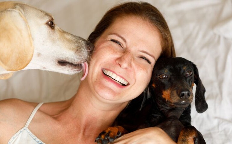 Jak pies okazuje miłość? Na zdjęciu biały pies liże twarz kobiety, a czarny się przytula.